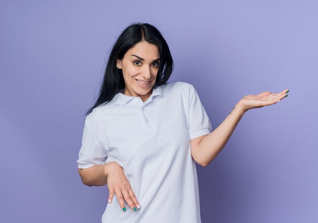 Sonriente joven morena caucásica sostiene la mano abierta aislada en la pared púrpura