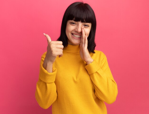 Sonriente joven morena caucásica sostiene la mano cerca de la boca y los pulgares para arriba en rosa