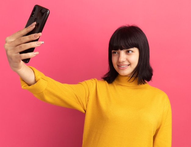 Foto gratuita sonriente joven morena caucásica sostiene y mira el teléfono tomando selfie en rosa