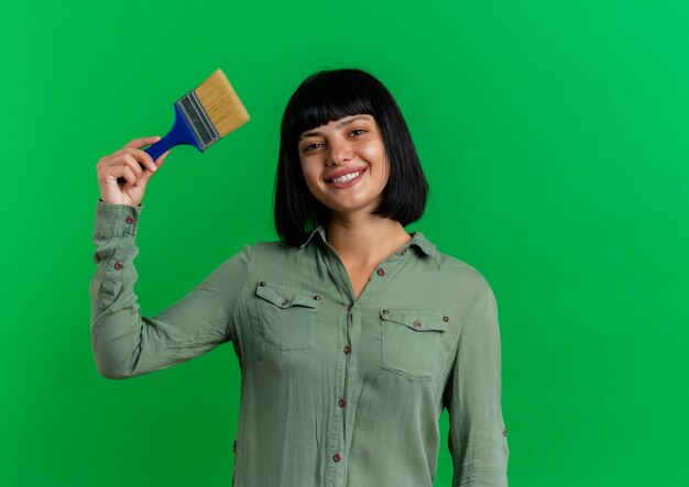 Sonriente joven morena caucásica sostiene pincel aislado sobre fondo verde con espacio de copia