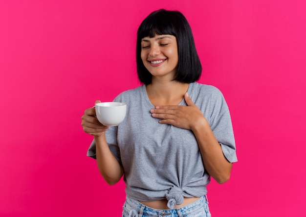 Foto gratuita sonriente joven morena caucásica sostiene la taza y pone la mano en el pecho