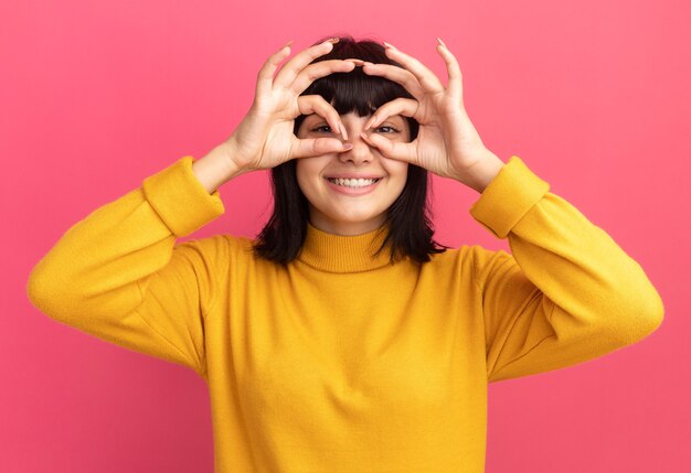 Sonriente joven morena caucásica a través de los dedos aislados en la pared rosa con espacio de copia