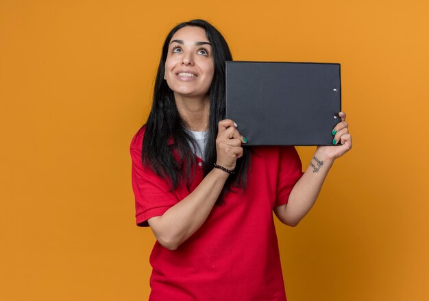 Sonriente joven morena caucásica vestida con camisa roja sostiene el portapapeles mirando hacia arriba aislado en la pared naranja