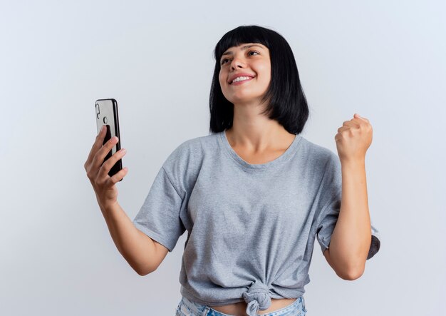 Sonriente joven morena mujer caucásica mantiene el puño y sostiene el teléfono mirando hacia arriba