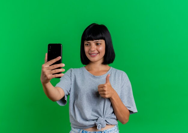 Sonriente joven morena mujer caucásica Thumbs up y mira el teléfono