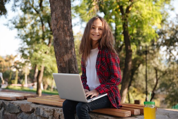 Sonriente joven morena sentada en el banco con la computadora portátil
