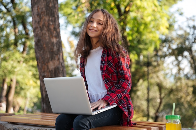 Sonriente joven morena sentada en el parque con ordenador portátil