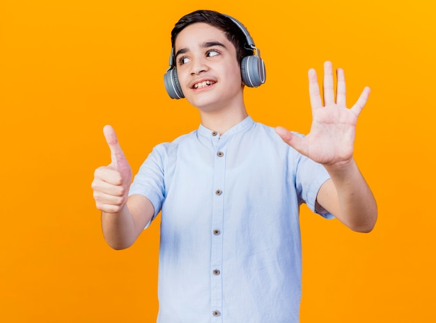 Sonriente joven muchacho caucásico con audífonos mirando al lado mostrando seis con manos aisladas sobre fondo naranja