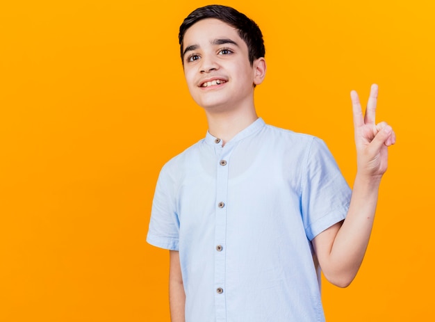 Sonriente joven muchacho caucásico mirando al lado haciendo el signo de la paz aislado sobre fondo naranja con espacio de copia