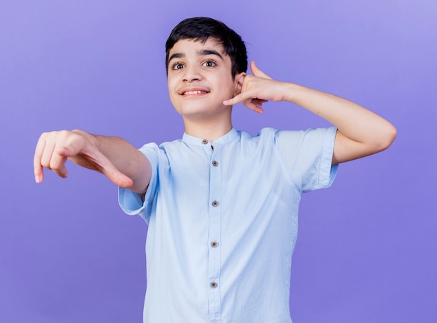 Sonriente joven muchacho caucásico mirando y apuntando directamente haciendo gesto de llamada aislado en la pared púrpura