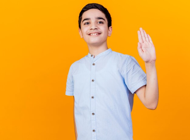 Sonriente joven muchacho caucásico mirando a la cámara haciendo hola gesto mirando a cámara aislada sobre fondo naranja con espacio de copia