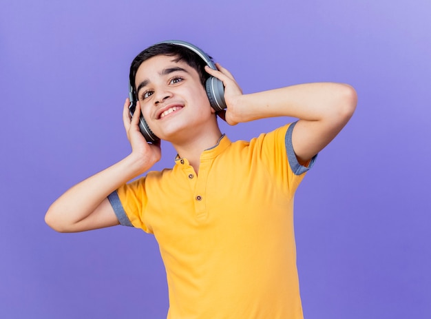 Sonriente joven muchacho caucásico mirando a la cámara tocando auriculares aislados sobre fondo púrpura