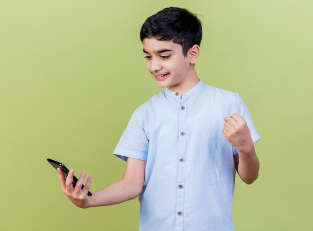 Foto gratuita sonriente joven muchacho caucásico sosteniendo y mirando el teléfono móvil haciendo sí gesto aislado sobre fondo verde oliva