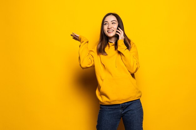 Sonriente joven mujer asiática casual hablando teléfono inteligente aislado sobre pared amarilla