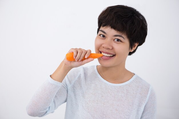 Sonriente joven mujer asiática mordiendo la zanahoria