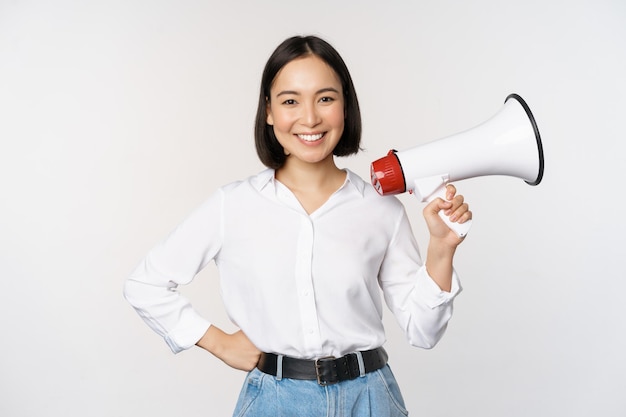 Sonriente joven mujer asiática posando con megáfono concepto de anuncio de noticias e información de pie sobre fondo blanco.