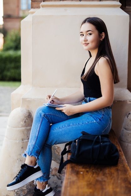 Sonriente joven mujer asiática sentada en un banco en el parque, tomando notas en un bloc de notas