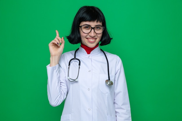 Sonriente joven mujer bastante caucásica con gafas en uniforme médico con estetoscopio apuntando hacia arriba