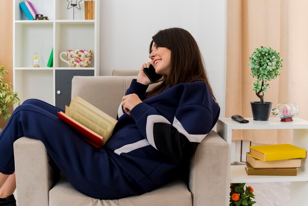 Sonriente joven mujer bastante caucásica sentada en un sillón en la sala de estar diseñada mirando directamente hablando por teléfono con el libro en las piernas