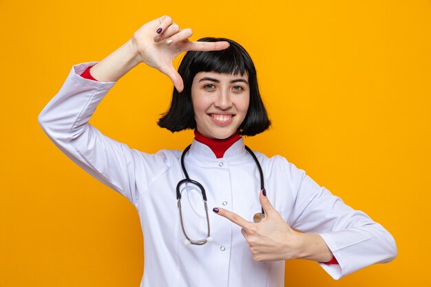 Sonriente a joven mujer bastante caucásica en uniforme médico con estetoscopio gesticulando el marco con las manos