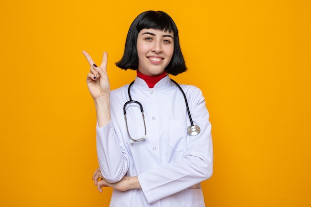 Sonriente joven mujer bastante caucásica en uniforme médico con estetoscopio gesticulando el signo de la victoria