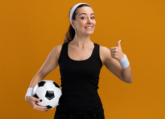Sonriente joven mujer bastante deportiva con diadema y muñequeras sosteniendo un balón de fútbol mirando al frente mostrando el pulgar hacia arriba aislado en la pared naranja