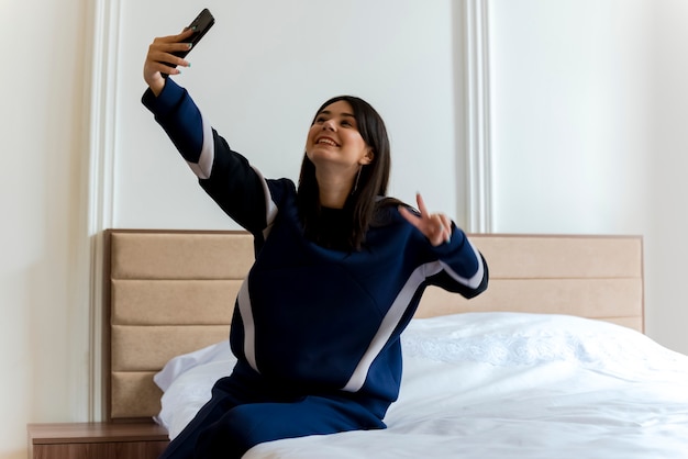 Sonriente joven mujer bonita caucásica sentada en la cama en el dormitorio haciendo el signo de la paz y tomando selfie