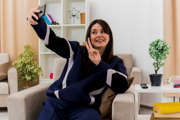 Sonriente joven mujer bonita caucásica sentada en un sillón en la sala de estar diseñada haciendo el signo de la paz y tomando selfie