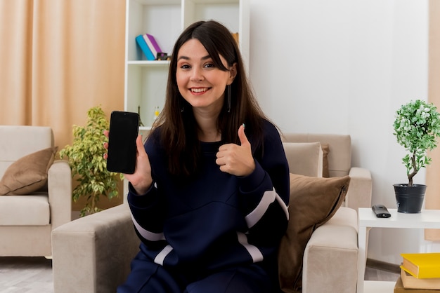 Sonriente joven mujer bonita caucásica sentada en un sillón en la sala de estar diseñada mostrando el teléfono móvil y mostrando el pulgar hacia arriba