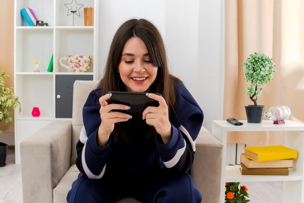 Sonriente joven mujer bonita caucásica sentada en un sillón en la sala de estar diseñada usando su teléfono móvil