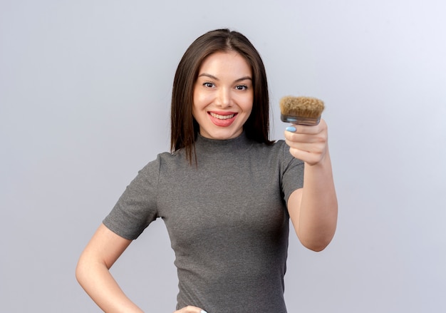 Sonriente joven mujer bonita estirando el pincel en la cámara y poniendo la mano en la cintura aislada sobre fondo blanco con espacio de copia