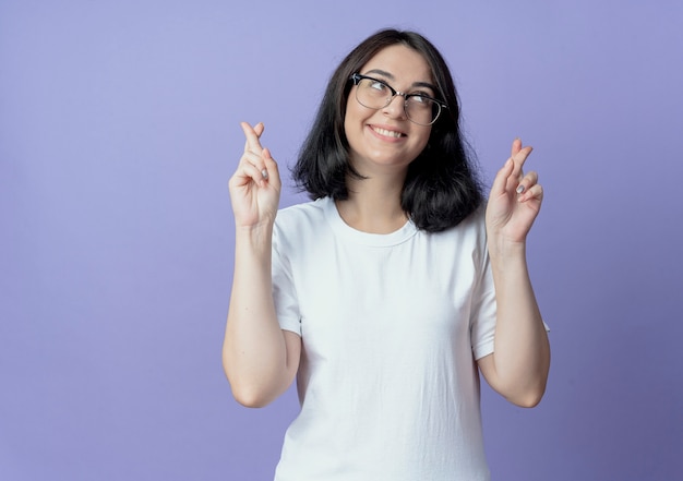 Sonriente joven mujer bonita con gafas mirando hacia arriba y haciendo gestos con los dedos cruzados