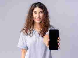 Foto gratuita sonriente joven mujer bonita mirando al frente estirando el teléfono móvil hacia el frente aislado en la pared blanca