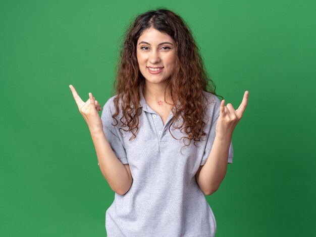 Sonriente joven mujer bonita mirando al frente haciendo signo de rock aislado en la pared verde