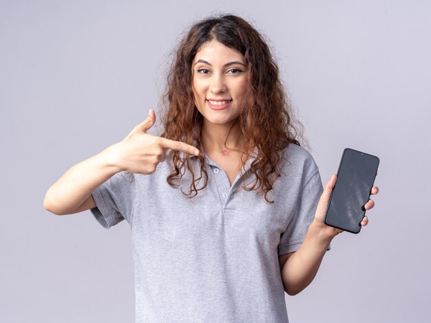 Sonriente joven mujer bonita mirando al frente mostrando el teléfono móvil al frente apuntando a él aislado en la pared blanca