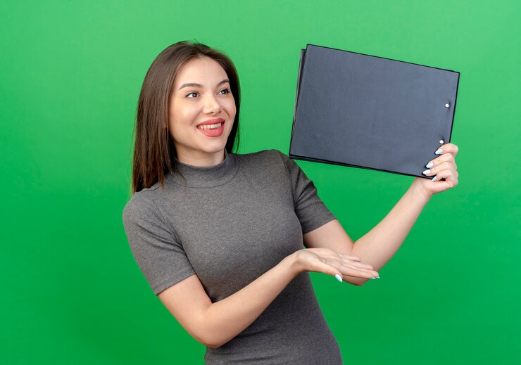 Foto gratuito sonriente joven mujer bonita mirando al lado sosteniendo y apuntando con la mano al portapapeles aislado sobre fondo verde