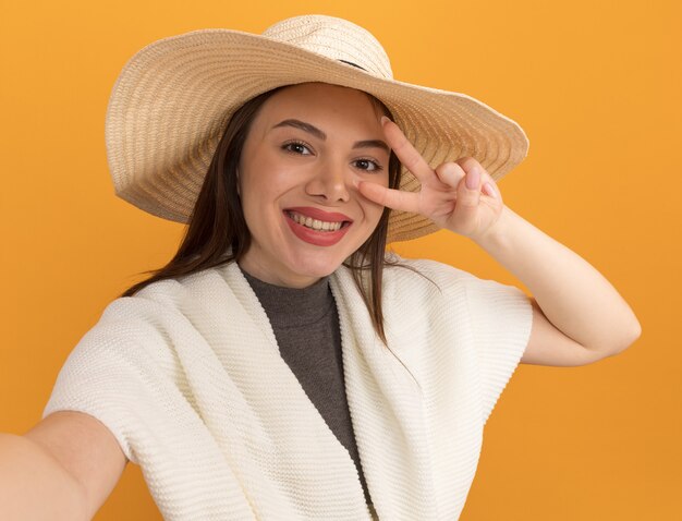 Sonriente joven mujer bonita con sombrero de playa mirando al frente estirando la mano hacia el frente mostrando el símbolo de signo v cerca del ojo aislado en la pared naranja