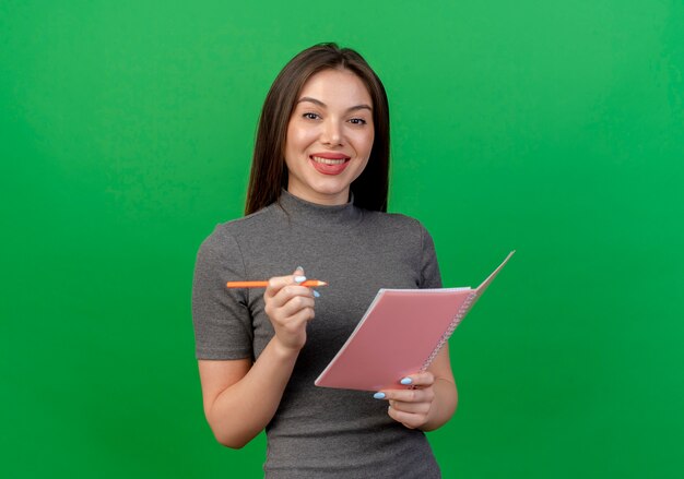 Sonriente joven mujer bonita sosteniendo bloc de notas y bolígrafo aislado en verde con espacio de copia