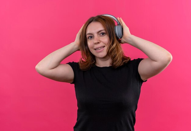 Sonriente joven mujer casual usando audífonos y poniendo las manos sobre ellos en el espacio rosa aislado