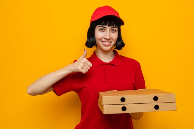 Sonriente joven mujer caucásica de entrega sosteniendo cajas de pizza y pulgar hacia arriba