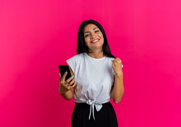 Sonriente joven mujer caucásica sostiene el teléfono y mantiene el puño