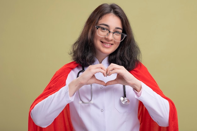 Sonriente joven mujer caucásica de superhéroe en capa roja vistiendo uniforme médico y estetoscopio con gafas haciendo señal de corazón