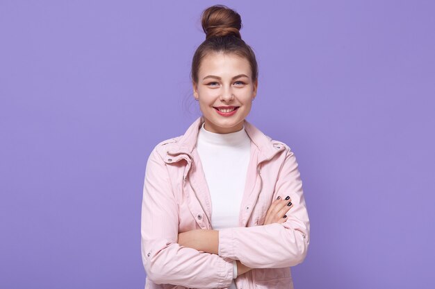 Sonriente joven mujer con chaqueta rosa pálido y camisa blanca posando aislada en la pared lila, manteniendo las manos cruzadas, mujer, expresando emociones positivas.