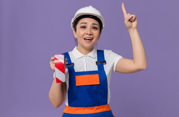 Sonriente joven mujer constructora asiática con casco de seguridad blanco sosteniendo cinta de advertencia y apuntando hacia arriba