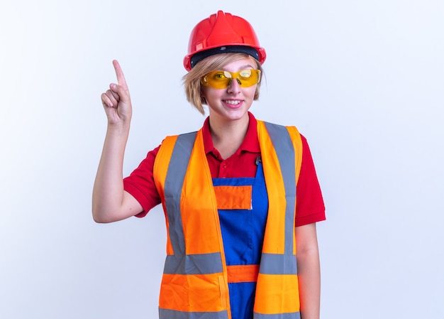 Sonriente joven mujer constructora en uniforme con gafas apunta hacia arriba aislado en la pared blanca