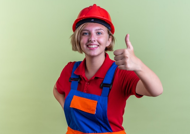 Sonriente joven mujer constructora en uniforme mostrando el pulgar hacia arriba aislado en la pared verde oliva