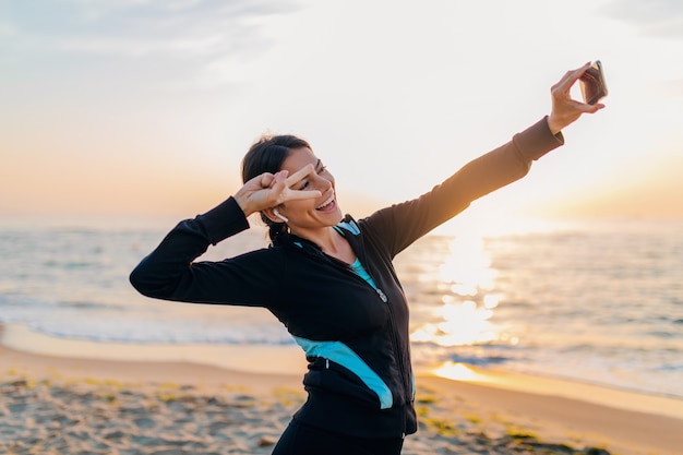 Sonriente joven mujer delgada atractiva haciendo ejercicios deportivos en la playa del amanecer de la mañana en ropa deportiva, estilo de vida saludable, escuchando música en auriculares, haciendo fotos selfie en el teléfono con humor positivo