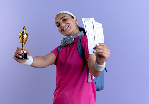 Sonriente joven mujer deportiva caucásica con diadema y muñequeras de bolsa trasera sostiene la copa ganadora y boletos aéreos aislados sobre fondo púrpura con espacio de copia