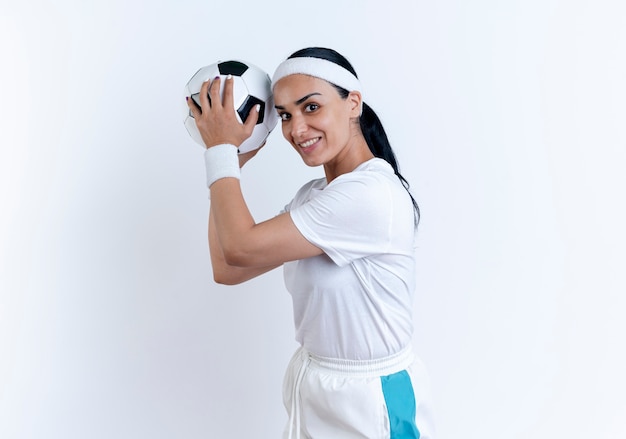 Sonriente joven mujer deportiva caucásica con diadema y muñequeras se encuentra de lado sosteniendo la bola aislada en el espacio en blanco con espacio de copia