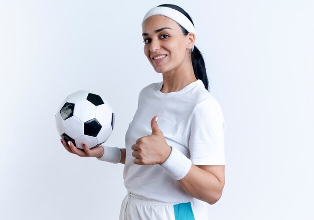 Sonriente joven mujer deportiva caucásica con diadema y muñequeras sostiene la bola y los pulgares para arriba de pie hacia los lados aislado en el espacio en blanco con espacio de copia